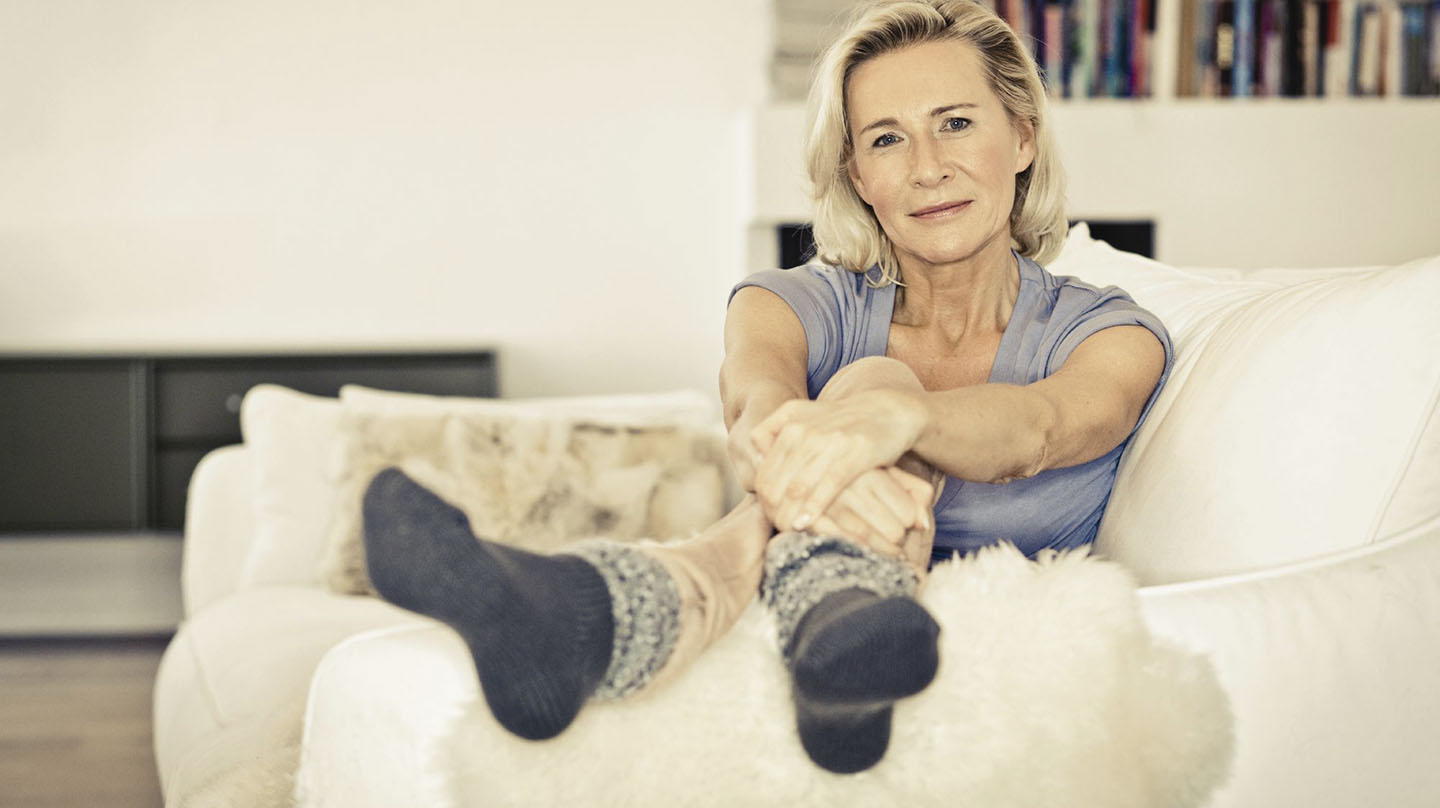 Hormone in den Wechseljahren: Lächelnde ältere Frau mit sitzt entspannt auf dem Sofa.