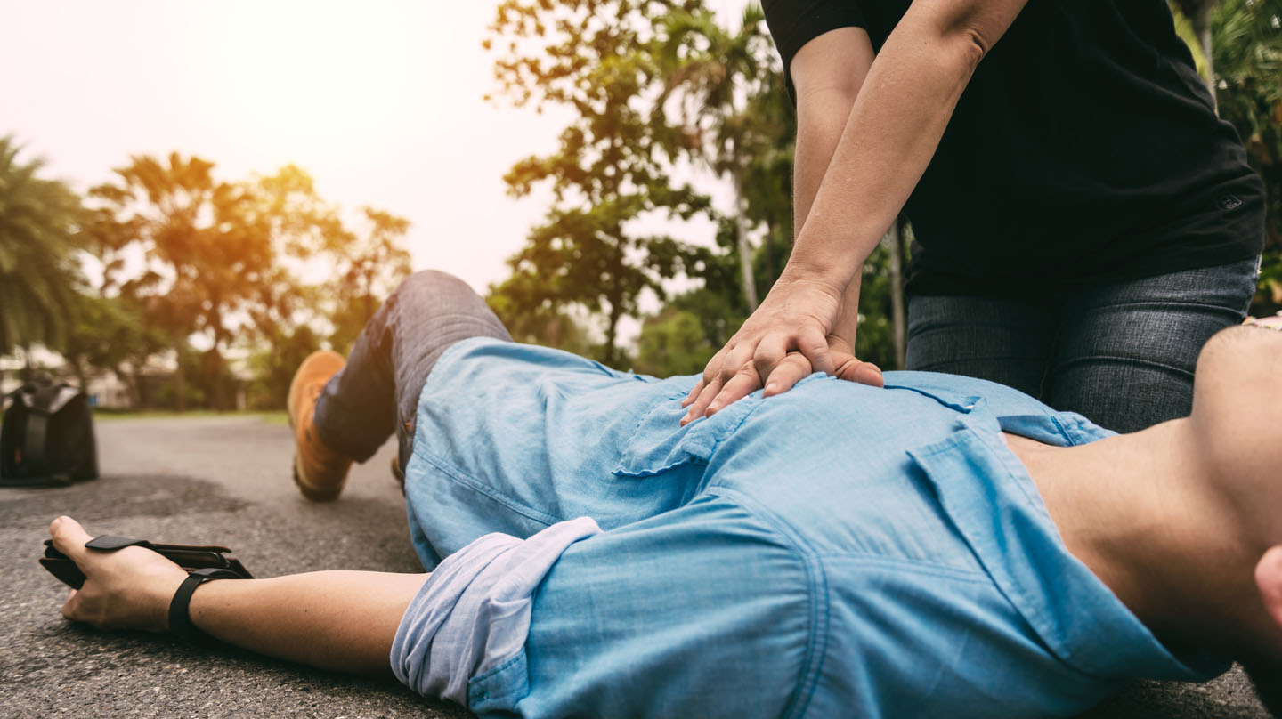 Erste Hilfe trotz Corona – das ist zu beachten Erste Hilfe: Junger Mann im blauen Hemd liegt auf der Straße. Ein anderer Mann macht mit den Händen Herzdruckmassage.